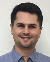 The image is a portrait of a smiling man with short hair, wearing a dark shirt and a tie, standing against a plain background.