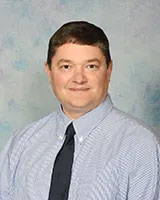 The image is a portrait of an adult male with short hair, wearing a blue shirt and tie, standing against a blue background.