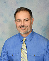 Man in a blue shirt and tie, smiling at the camera.