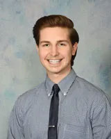 The image shows a young man with short hair, wearing a gray shirt and a dark tie. He has a light beard and is smiling at the camera. Behind him, there s a blurred background that appears to be an indoor setting with blue lighting.