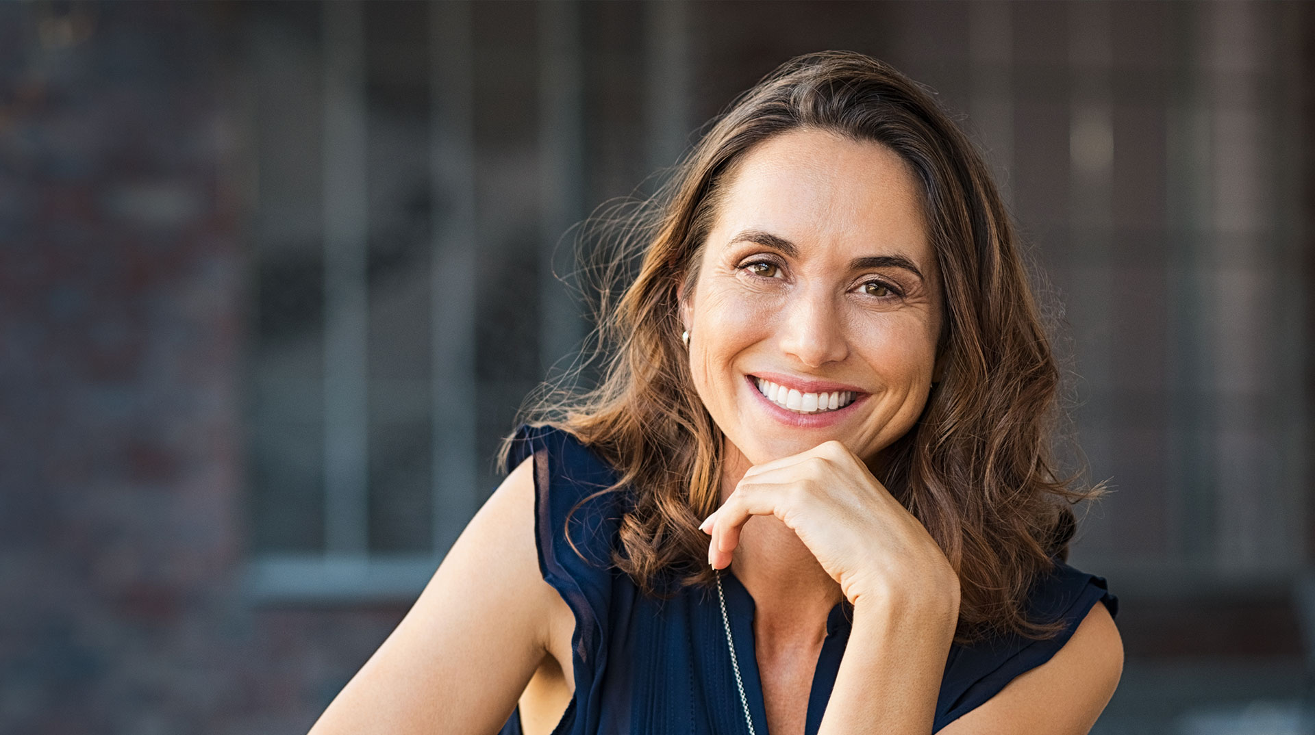 A woman with short hair is smiling and leaning forward slightly, posing for a portrait against a brick wall.