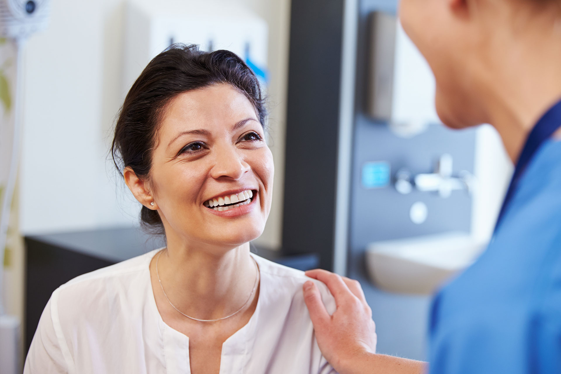 The image shows a woman in a dental office, smiling at another woman who is seated and appears to be receiving dental care.