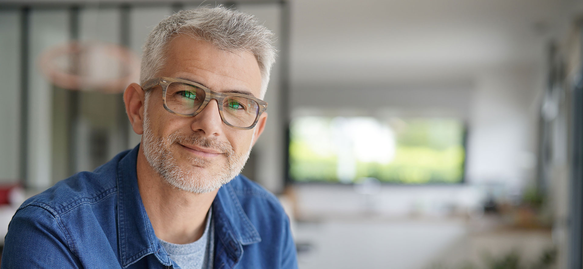 The image shows a man with gray hair, wearing glasses and a blue shirt, posing for the camera in an indoor setting.