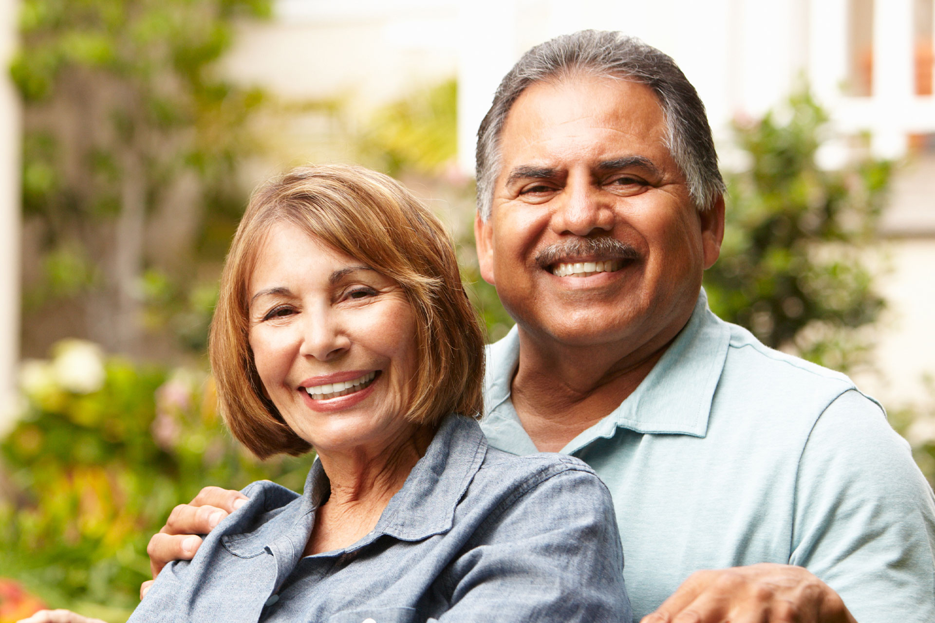 The image shows a man and a woman, both smiling and appearing to be in a happy mood. They are embracing each other, suggesting a close relationship or affection between them. The setting seems to be outdoors during the daytime, with a clear sky visible above them.