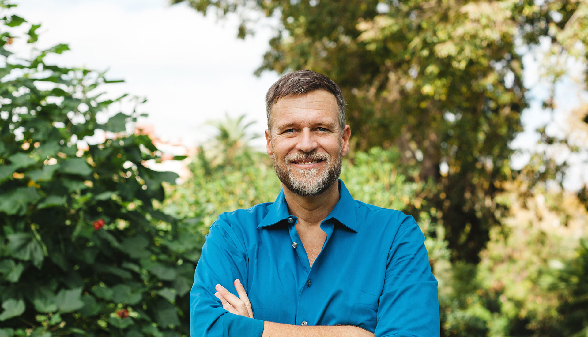 The image shows a man standing outdoors, posing for the camera with his arms crossed. He is wearing a blue shirt and has short hair. Behind him, there are plants and trees, suggesting a garden or park setting.