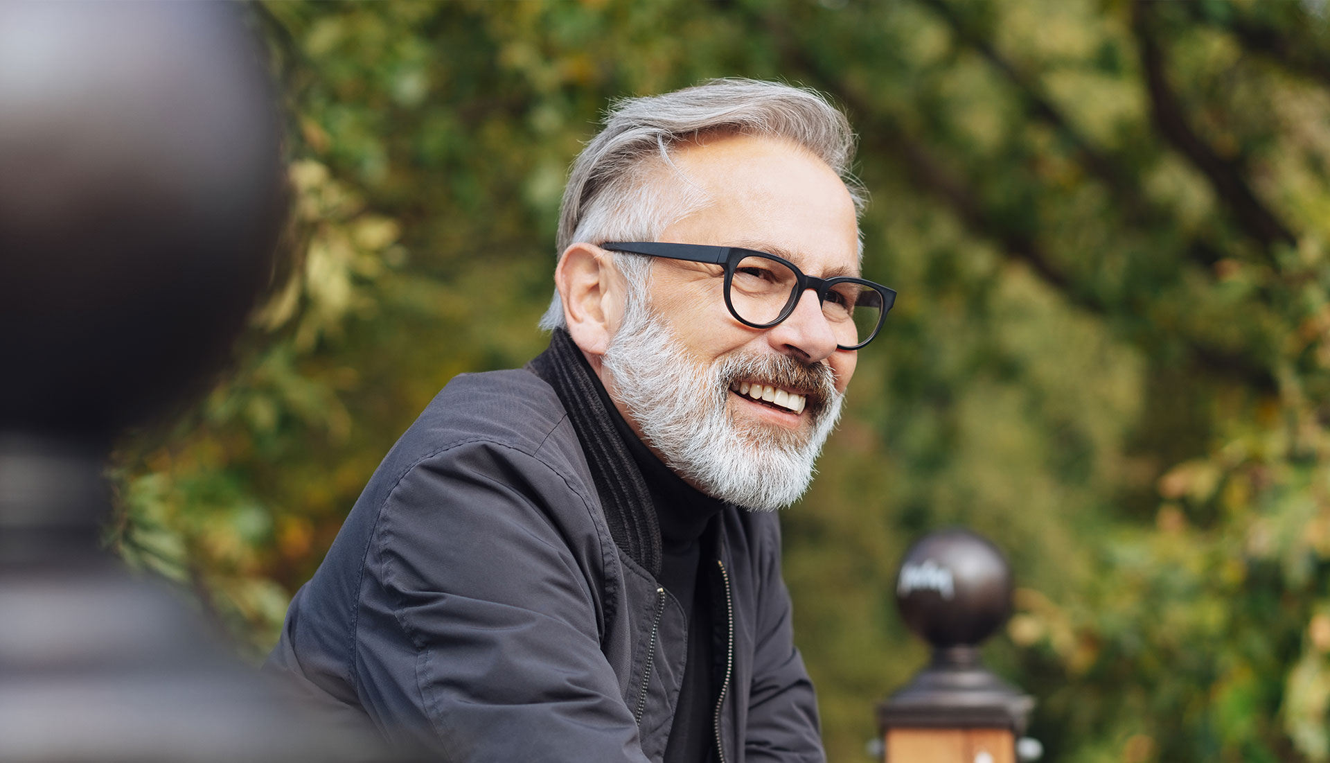 The image shows a man with a beard and glasses, wearing a dark jacket, smiling at the camera. He has short hair and appears to be in an outdoor setting during daylight.