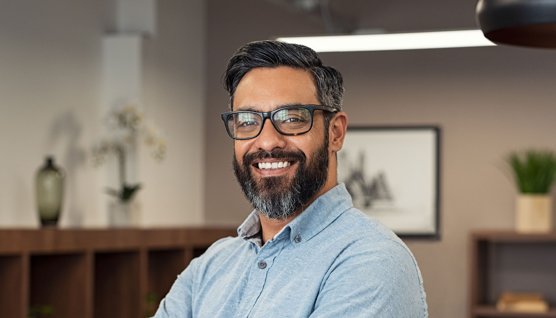 A man with a beard and glasses is smiling at the camera, standing in an office environment.