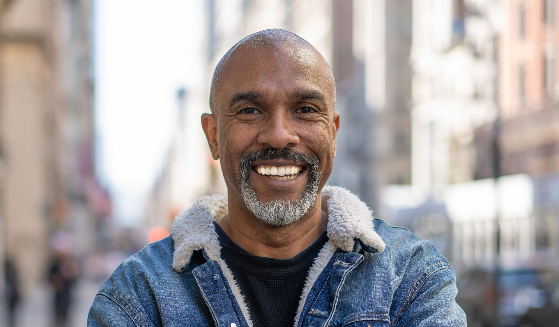 The image features a smiling man with a beard and mustache, wearing a denim jacket, standing on a city street.