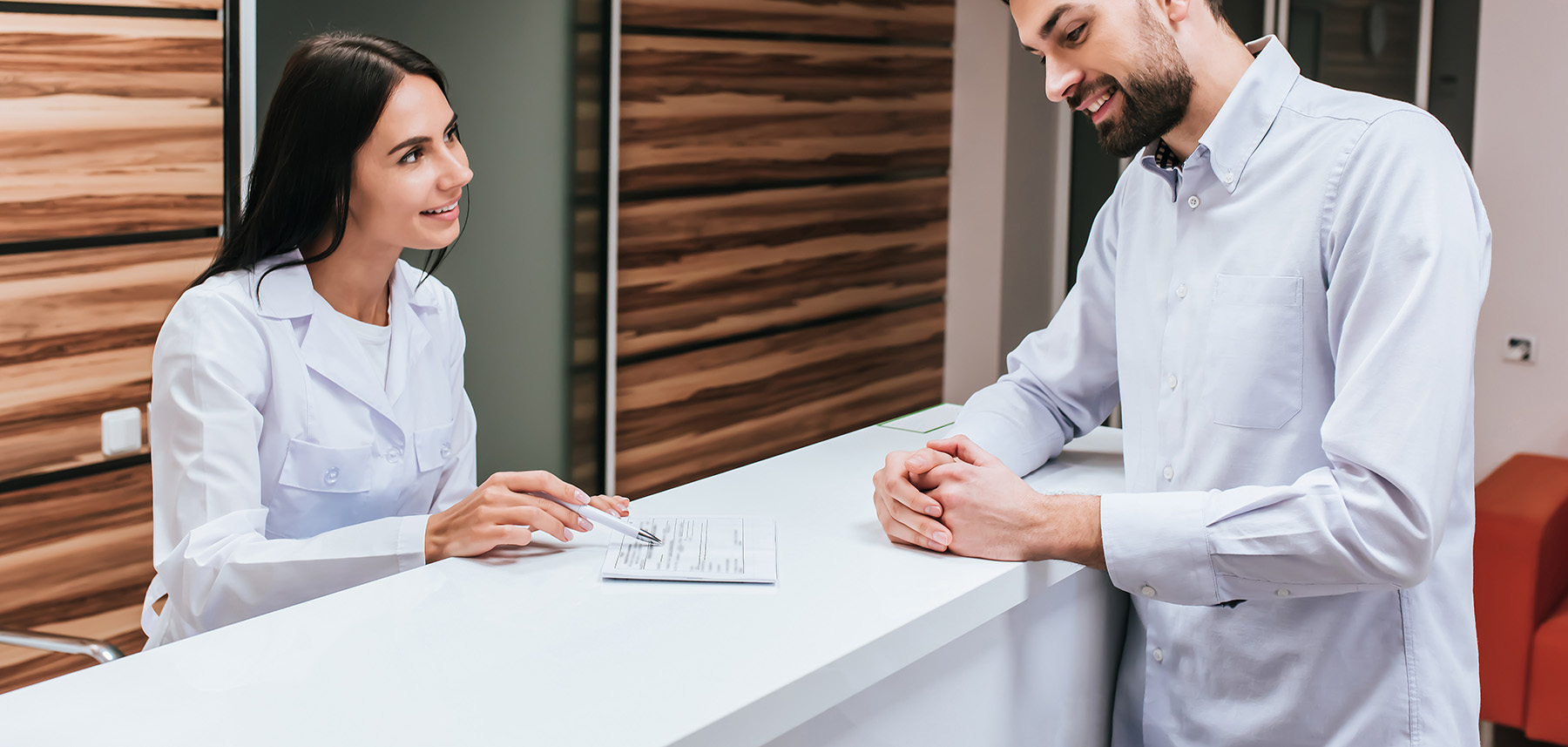 The image shows a man and a woman in professional attire, possibly in a medical or dental setting, with the man seated at a desk and the woman standing beside him.