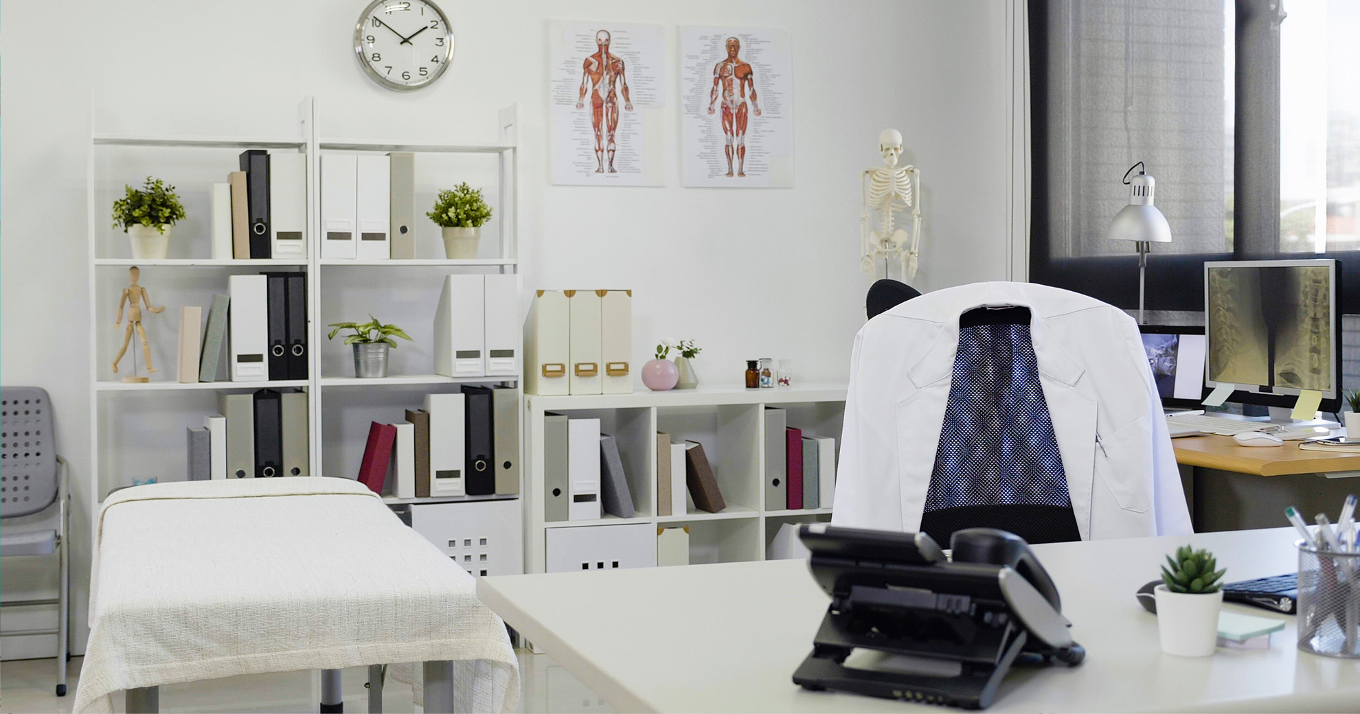 An office setting with medical-related equipment and furniture, including a desk, chair, and shelves filled with various items.