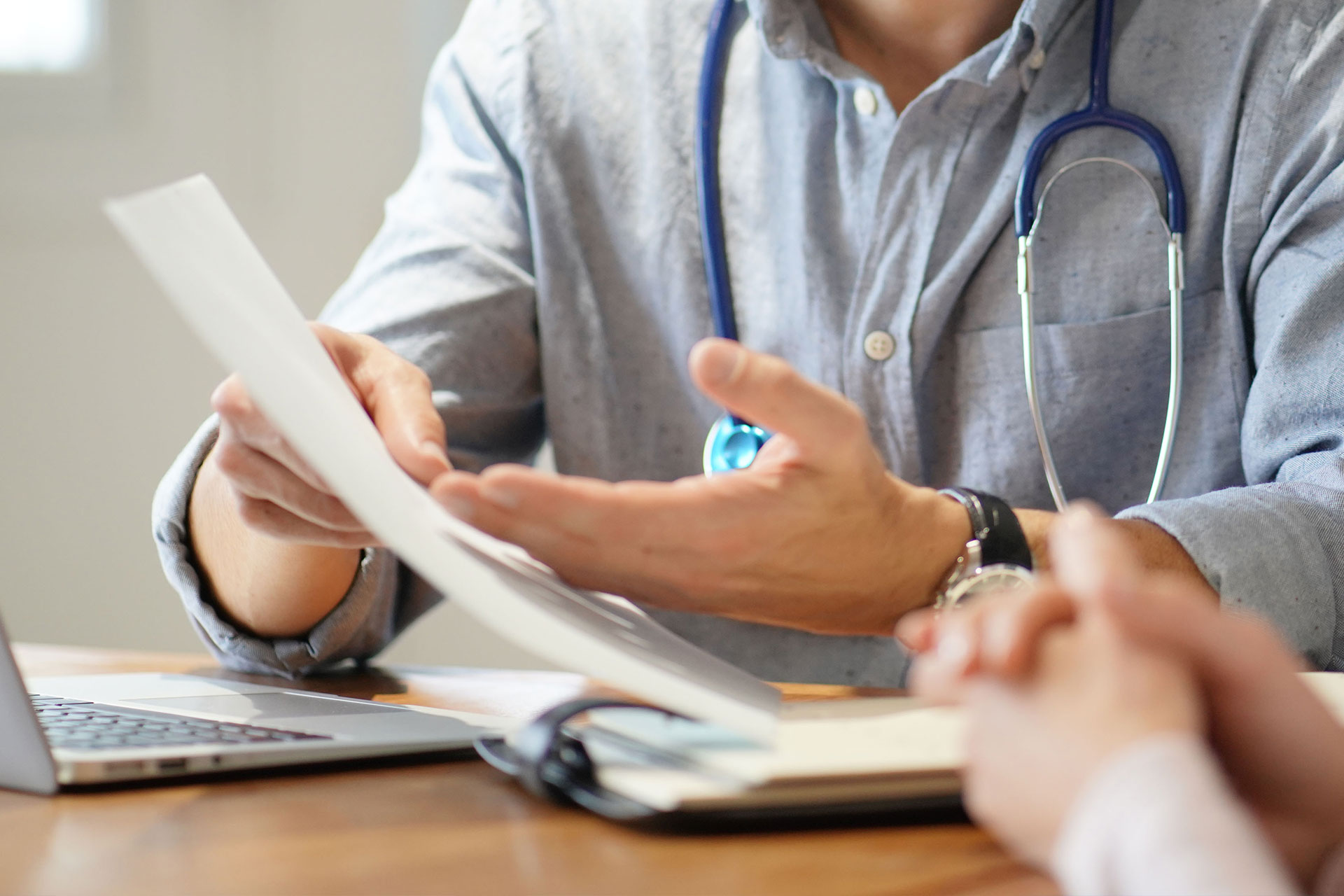 A man in a blue shirt and stethoscope is holding a document, looking at it with concentration while seated at a table.