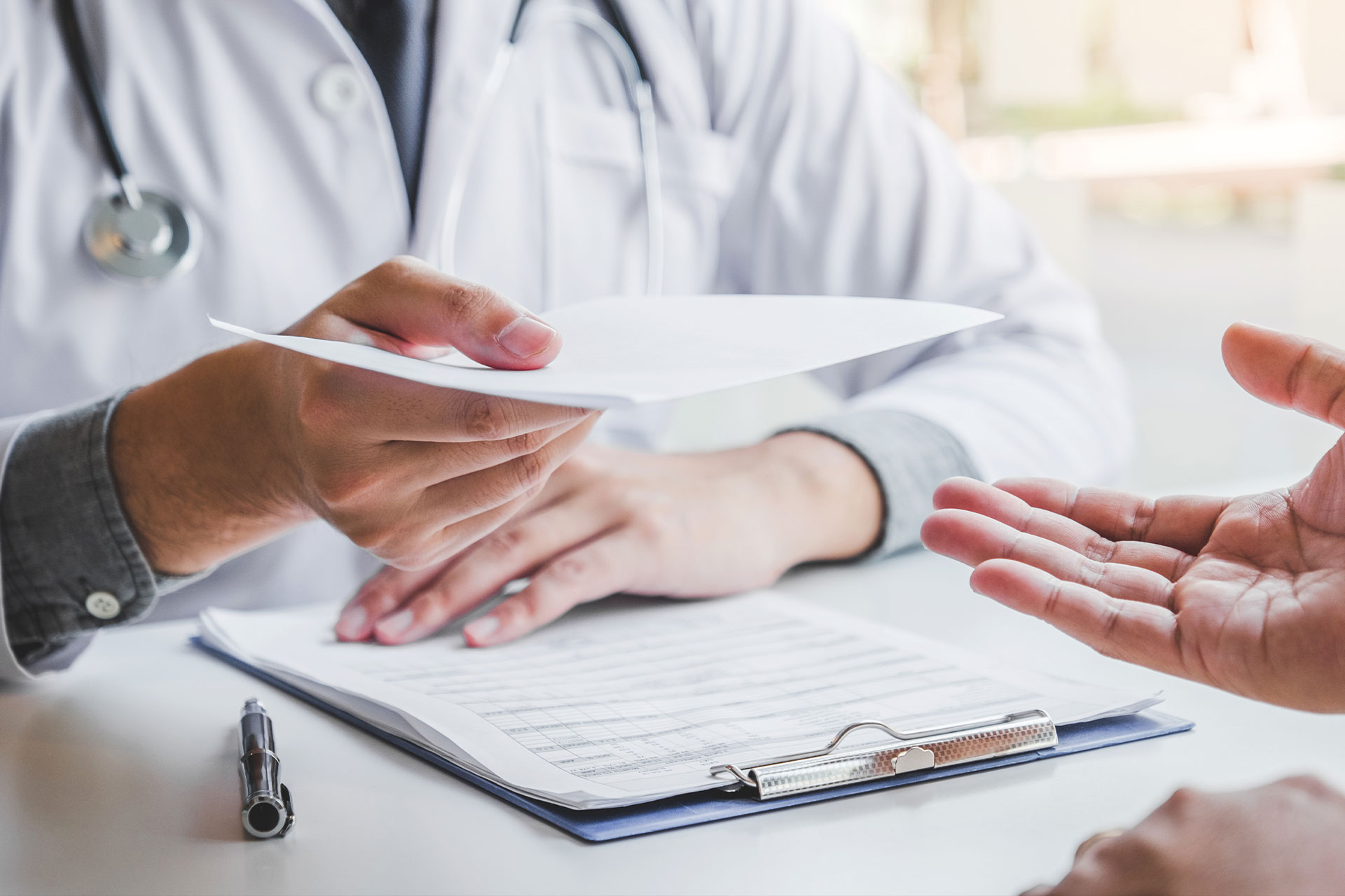 The image shows a medical professional, likely a doctor or nurse practitioner, seated at a desk with a patient s hands resting on the desk. The medical professional is holding a piece of paper and appears to be engaged in a conversation with the patient.