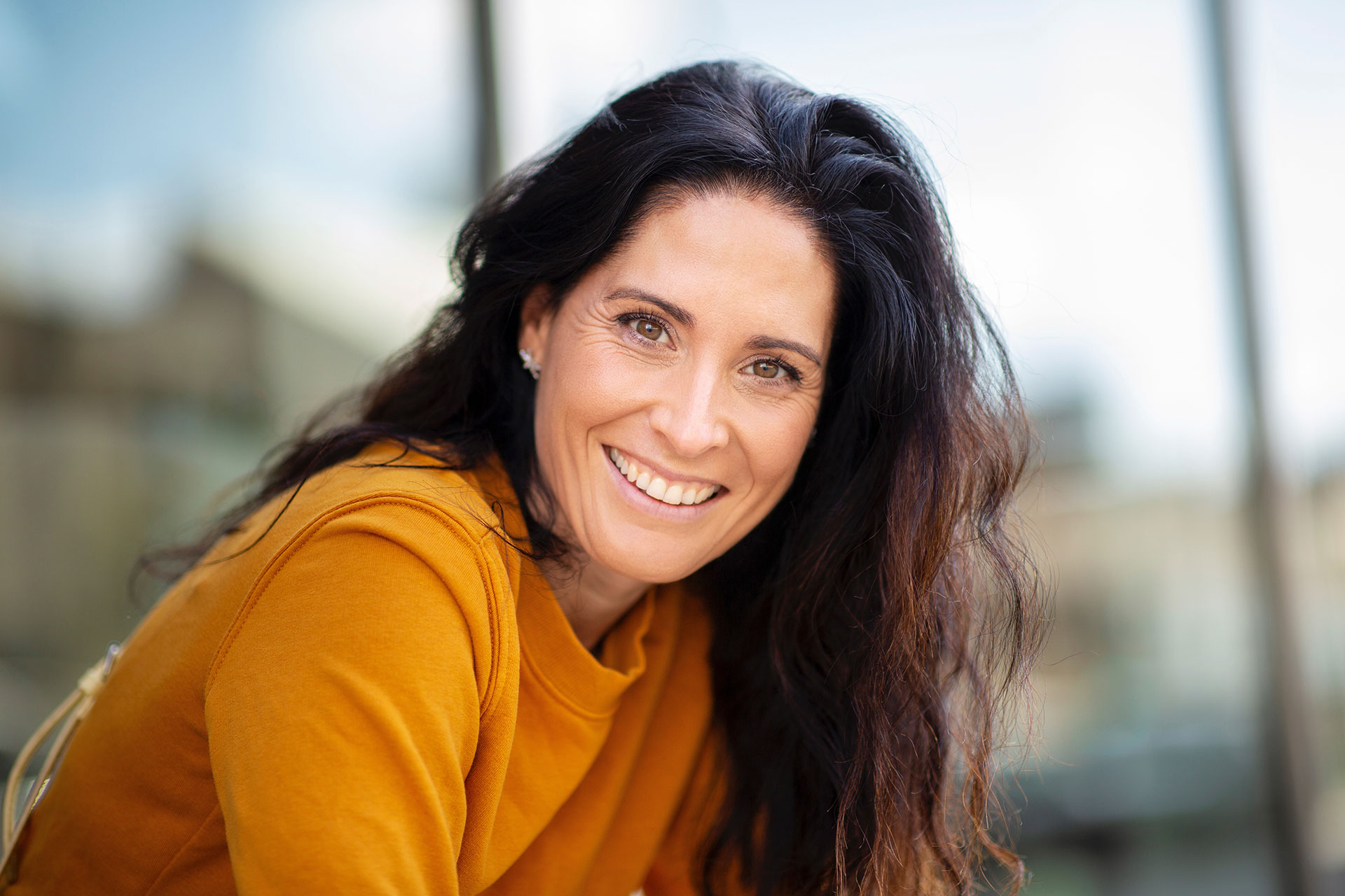 The image features a woman with long hair smiling at the camera, wearing a yellow top and standing outdoors.