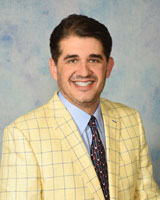 The image shows a man wearing a yellow blazer, standing against a blue background with a white wall on the left. He has dark hair and is smiling at the camera.