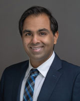 The image is a portrait of a man with a smile, wearing a suit and tie, against a neutral background.