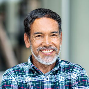 The image features a man with a beard and mustache, smiling at the camera. He is wearing a plaid shirt and appears to be indoors with a window in the background.